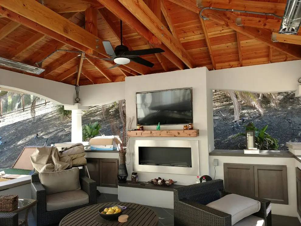 Solid roof pavilion interior with exposed wood beams, ceiling fan, infrared heaters, wall-mounted TV, linear fireplace, and seating, in a Danville backyard