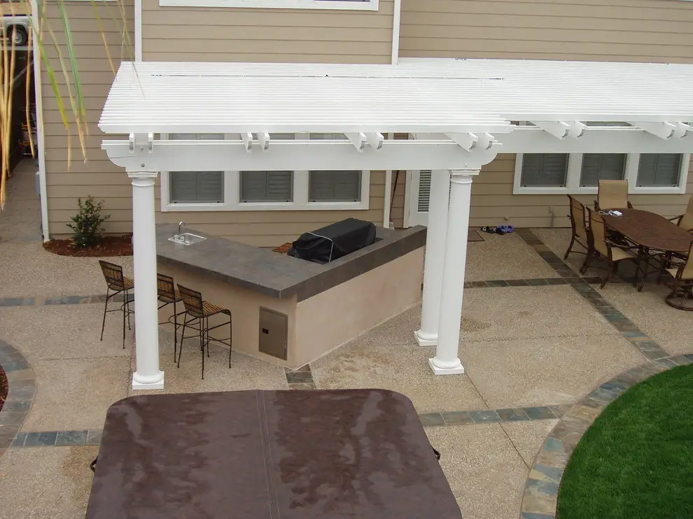 White pergola with round columns over a stucco built-in BBQ island with a tile countertop and bar stools, beside a covered hot tub