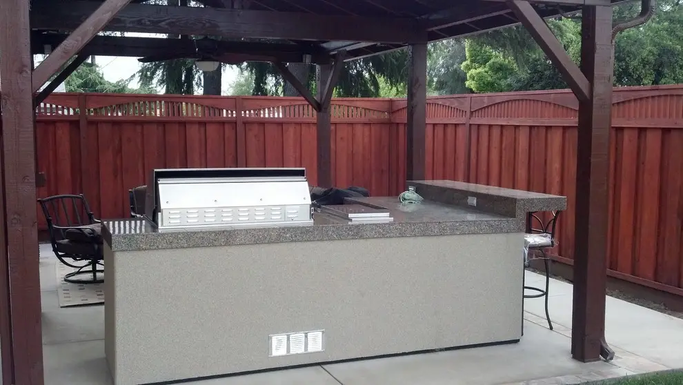 Built-in BBQ island with a stainless steel grill, granite countertop with a raised bar, and a stucco base, under a wood pavilion frame beside a redwood fence