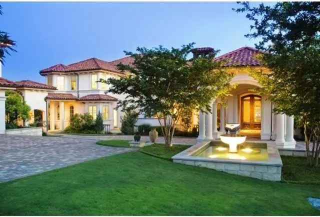 Stucco home with a clay tile roof and a stone fountain in front of the columned entry, seen from the lawn at dusk