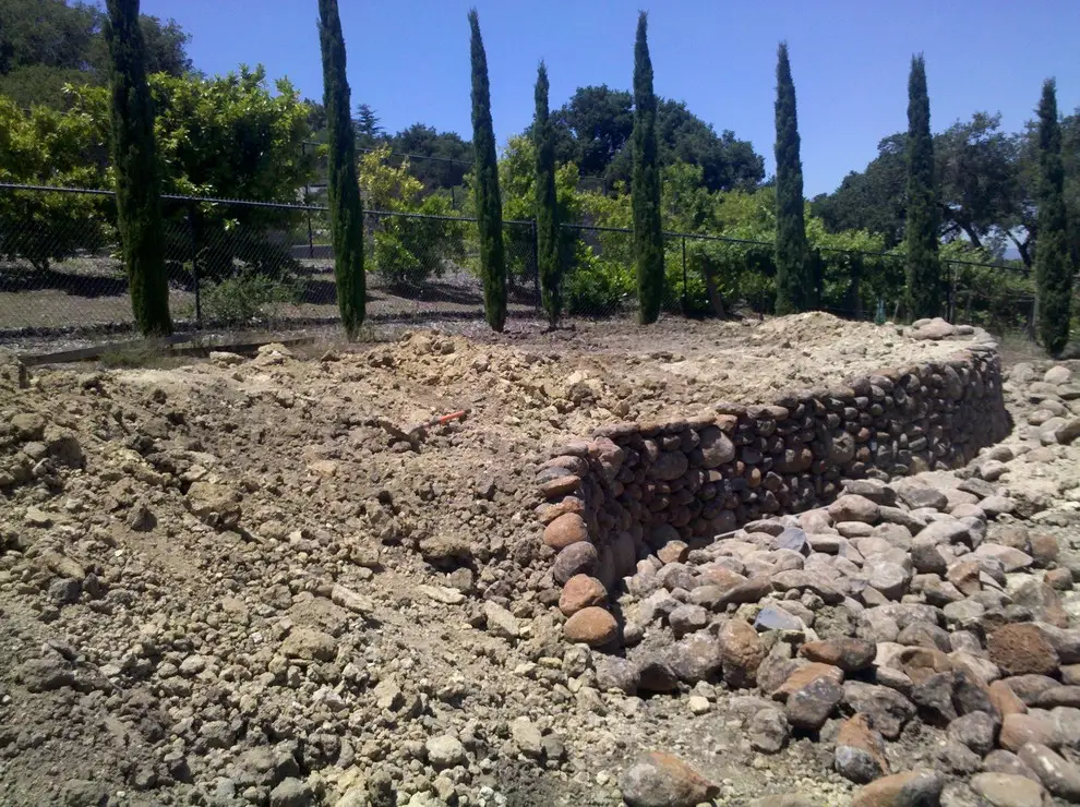 River rock retaining wall under construction, curving through freshly graded soil with tall cypress trees behind a chain-link fence