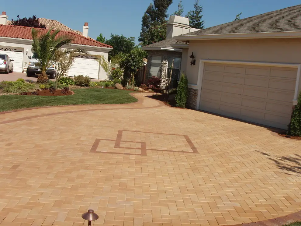 Curved herringbone paver driveway with a border band and geometric inlay, leading to a two-car garage beside a lawn and planting bed in Morgan Hill