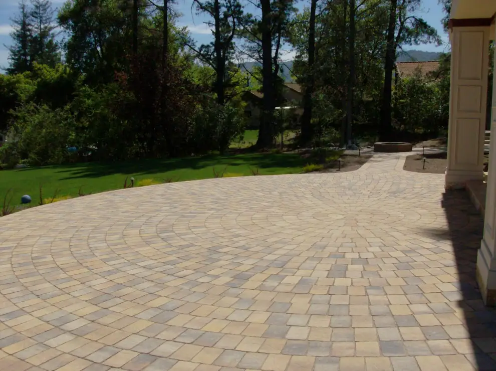 Circular paver patio with a radiating pattern beside a lawn, with path lights, a fire pit in the distance, and a column at the right edge
