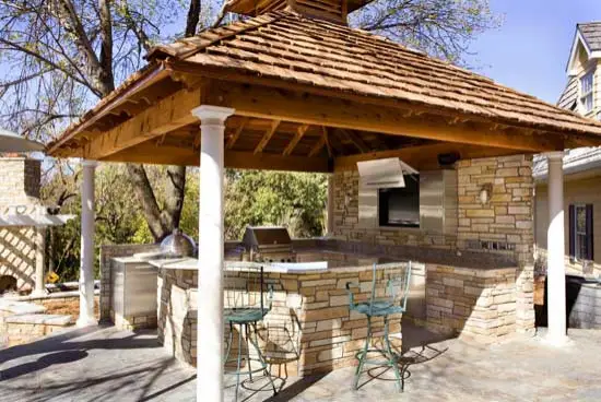 Cedar shake roof pavilion over a stacked stone outdoor kitchen island with a built-in grill and bar stools, with white columns on a flagstone patio