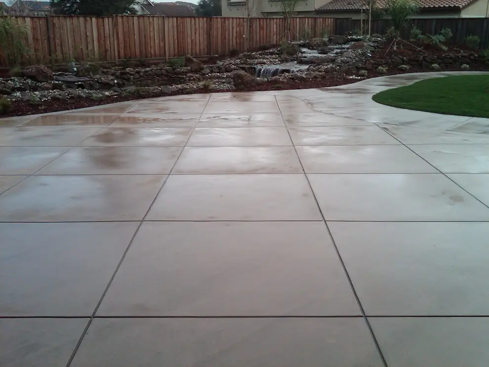 Wet scored concrete patio with a curved edge beside a lawn, with a rock-lined stream and waterfall in the planting bed behind