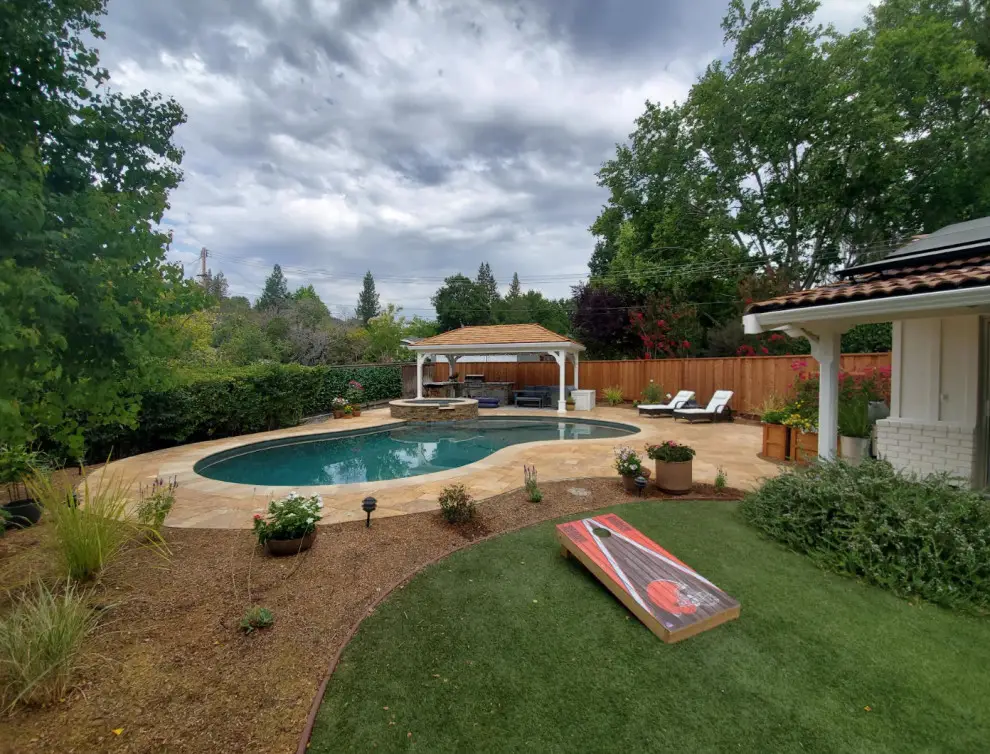 Wide backyard view of a kidney-shaped pool and spa with a solid roof pavilion, paver deck, mulch planting beds, and artificial turf lawn