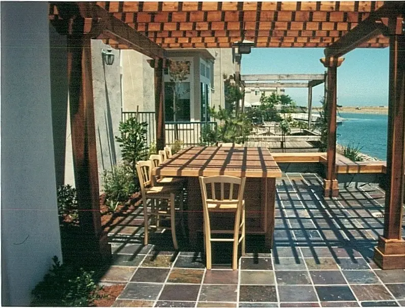 Wood pergola with a slatted roof over a tall table and bar chairs on a multicolor stone tile patio, with a waterfront view beyond