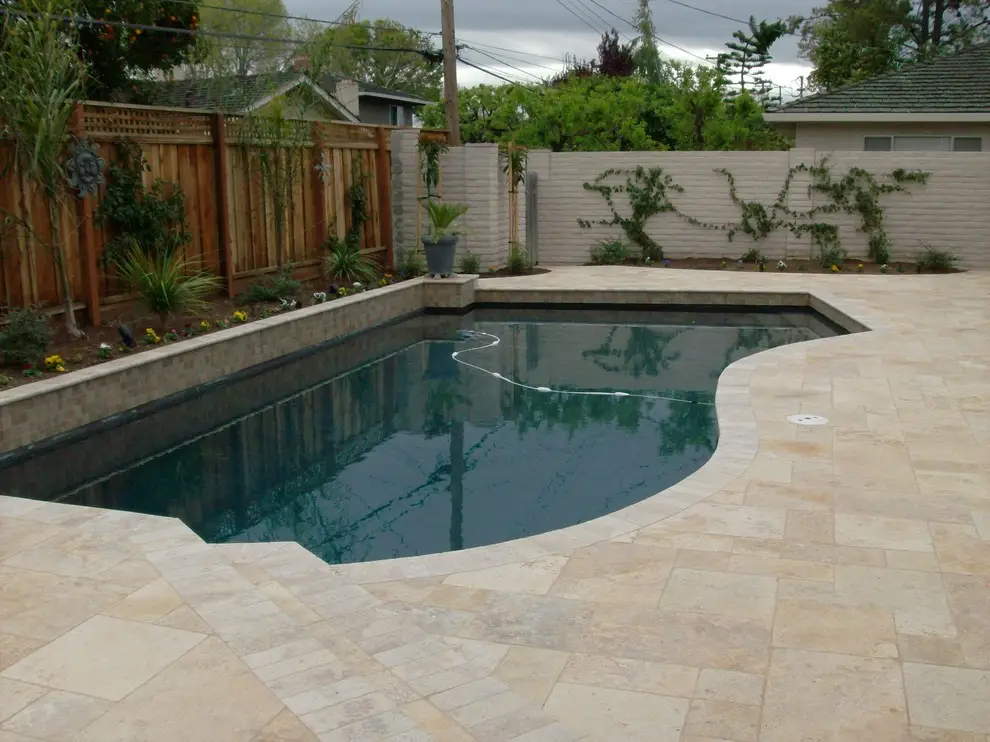 Freeform pool with a tile-faced raised wall and a light stone paver deck, beside a wood fence and a planting bed