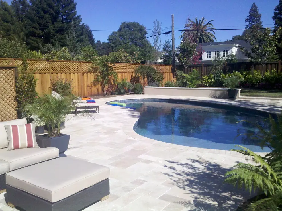 Curved pool with a light stone paver deck, an outdoor sectional with a striped pillow and an ottoman, and a lattice-topped wood fence behind