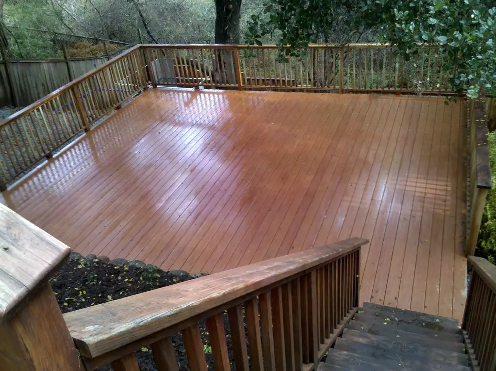 Large stained wood deck with a balustrade railing on three sides and a stairway in the foreground, above a mulched planting bed