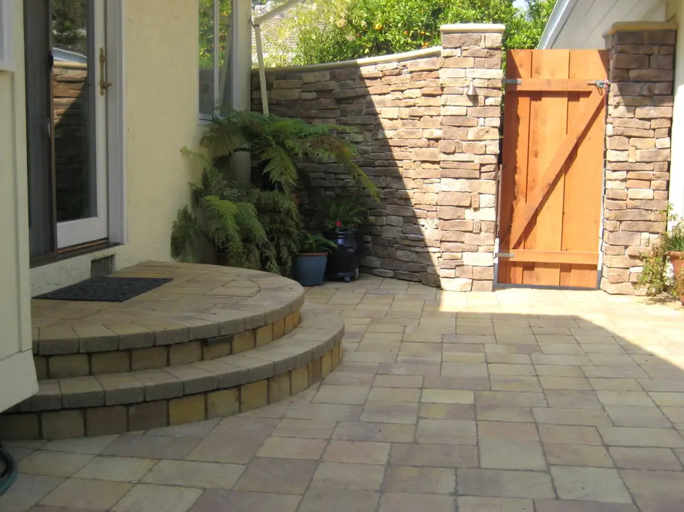 Paver courtyard with curved entry steps, a stacked stone wall, and a cedar gate beside potted ferns