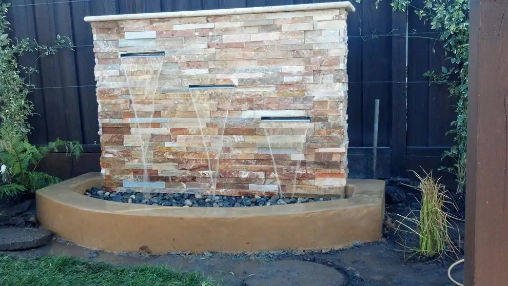 Stacked stone wall fountain with three sheer descent spouts over a curved stucco basin filled with dark river rock